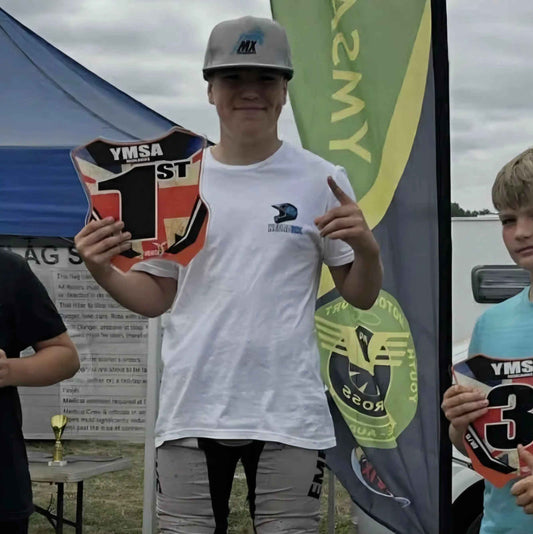 Motocross rider in casual apparel holding first place YMSA sign at outdoor event