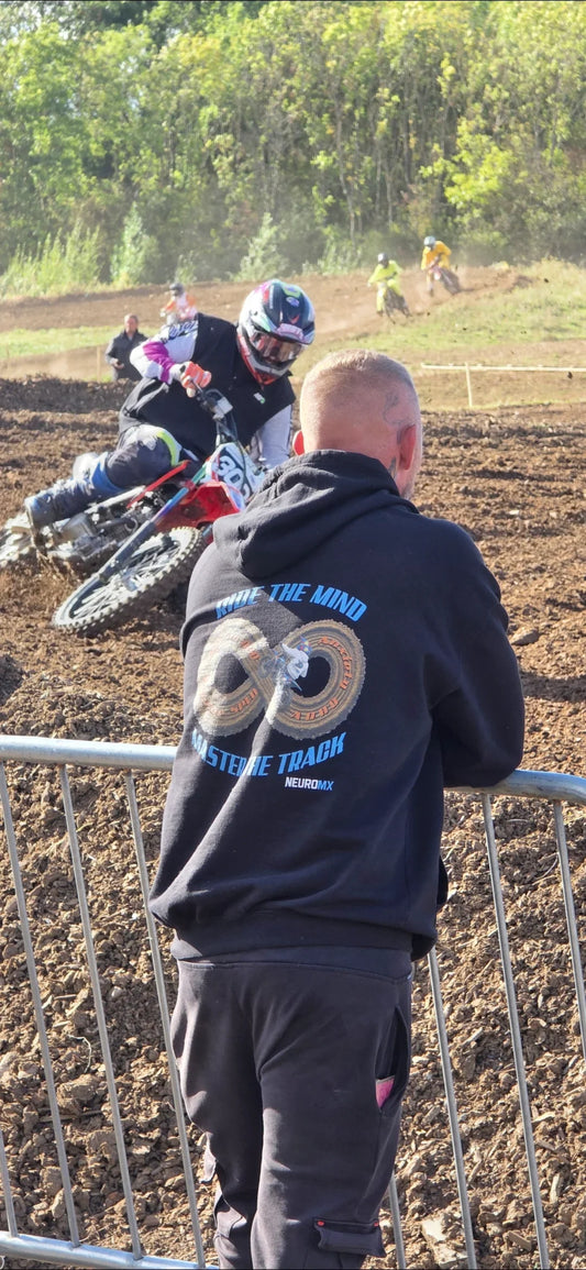 Motocross rider cornering on a dirt track, person in NeuroMX hoodie watches trackside.